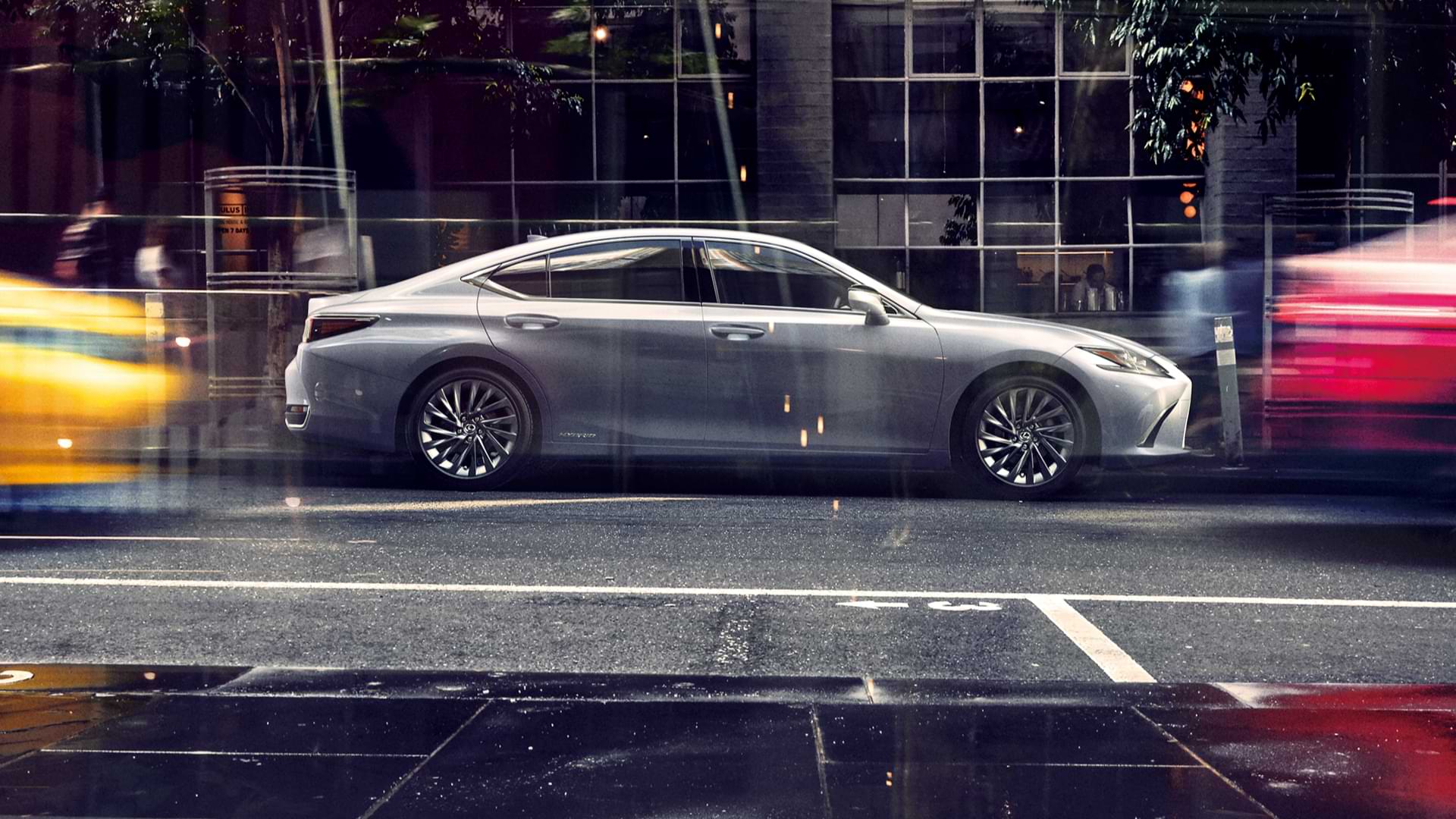 2021 ES 300h sits parked on a wet road. A red and yellow can be seen, blurred, flashing by.
