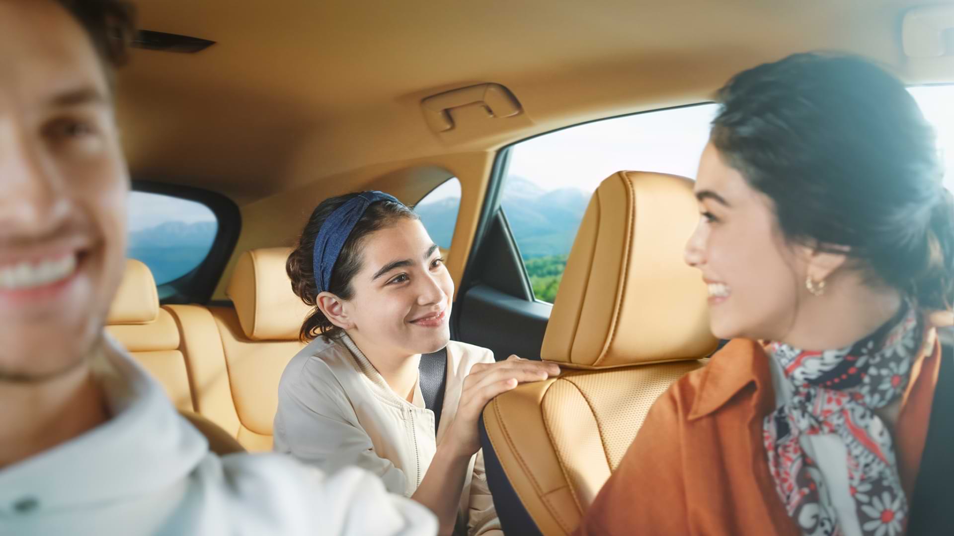 A woman sits in the front seat, a man driving. A child sits in the backseat, smiling at the woman, who is peering over the head rest into the backseat, smiling back at the child.