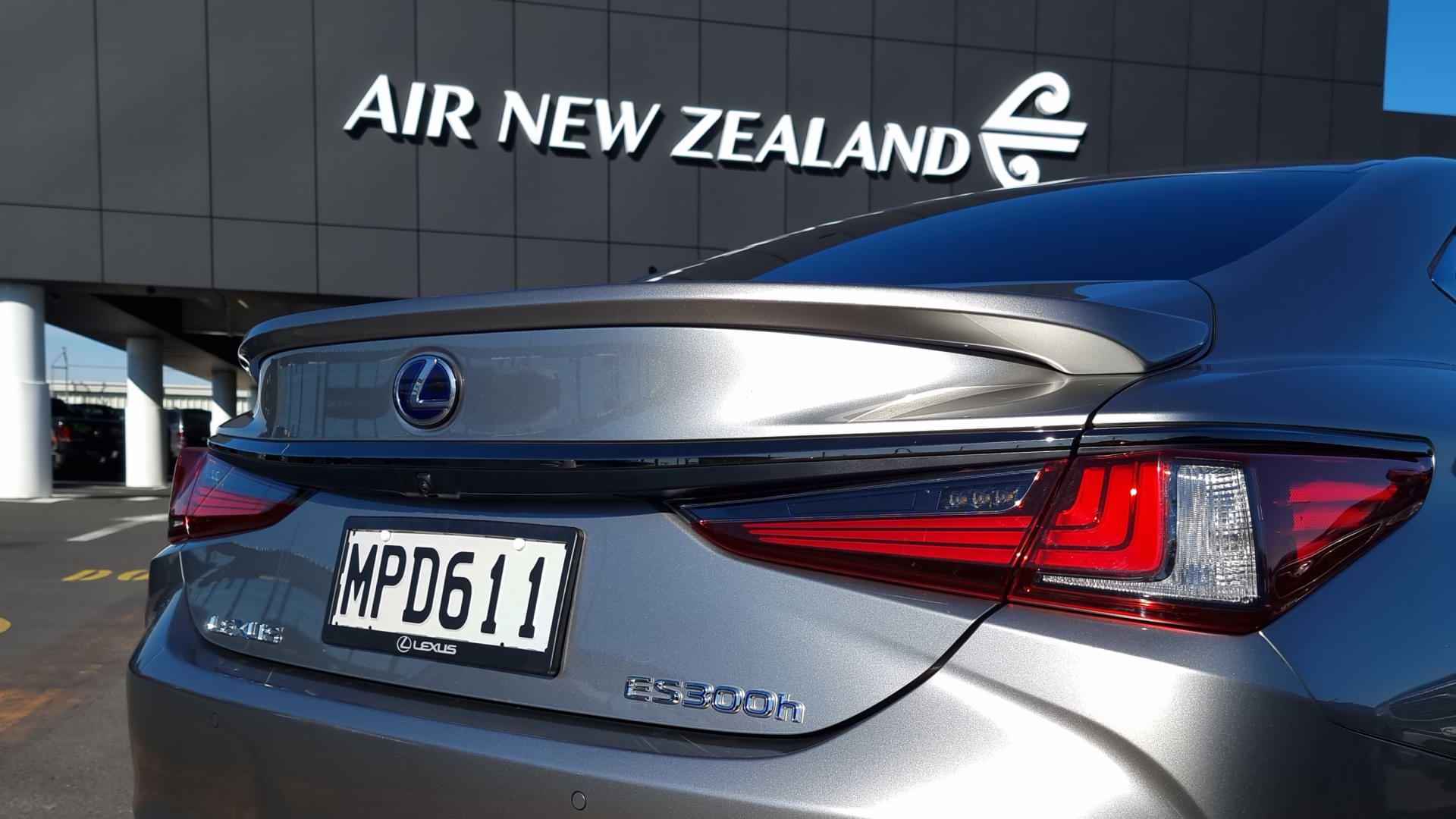 Silver Lexus ES 300h sits at the NZ airport carpark, with Air New Zealand terminal in the background.